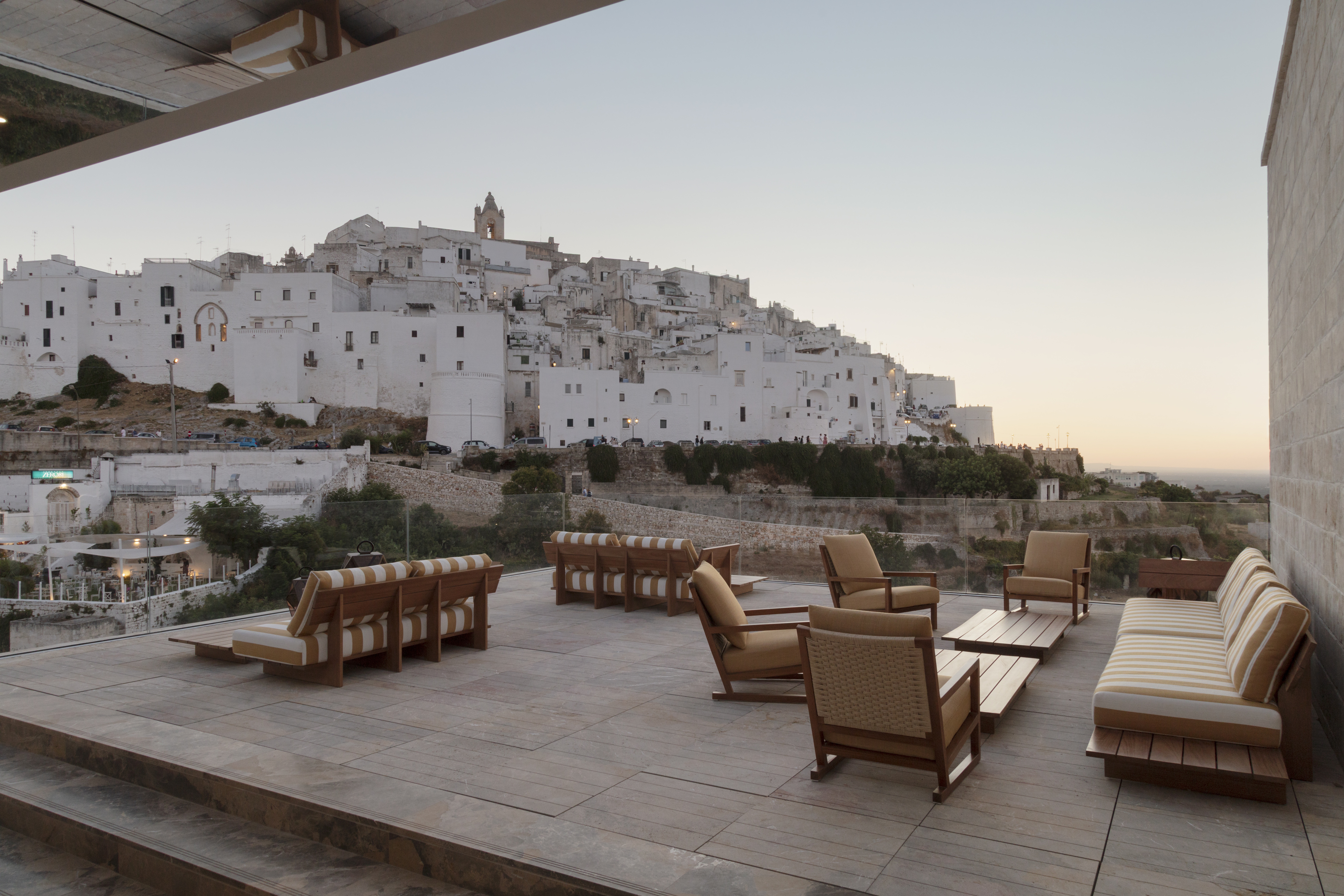 A picturesque southern Italian town with white-washed buildings overlooks a stylish terrace furnished with stripy furniture at dusk.