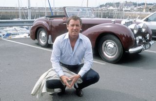 A promotional shot from the original 1980s Bergerac, showing John Nettles as Jim Bergerac, crouched in the car park at a marina in front of a cherry-red open top Triumph Roadster. Various yachts are just about visible in the background.