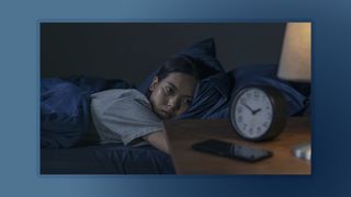 A woman lies in bed in the middle of the night unable to sleep, watching her alarm clock which is placed on a nightstand in the foreground of the image