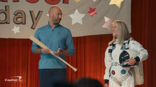 a man holding a baseball bat stands on a stage alonside a female astronaut in a white spacesuit