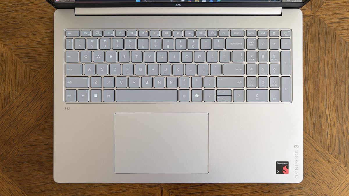 Top view of a silver laptop keyboard with a compact gray key layout and a touchpad, resting on a wooden tabletop. The laptop features &ldquo;OMNIBOOK 3&rdquo; and &ldquo;Snapdragon&rdquo; logos.