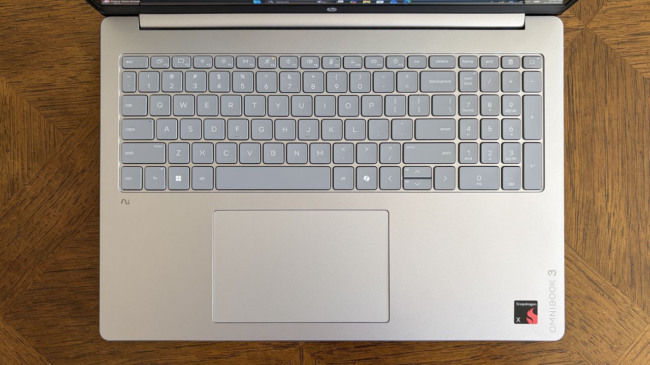 Top view of a silver laptop keyboard with a compact gray key layout and a touchpad, resting on a wooden tabletop. The laptop features &ldquo;OMNIBOOK 3&rdquo; and &ldquo;Snapdragon&rdquo; logos.