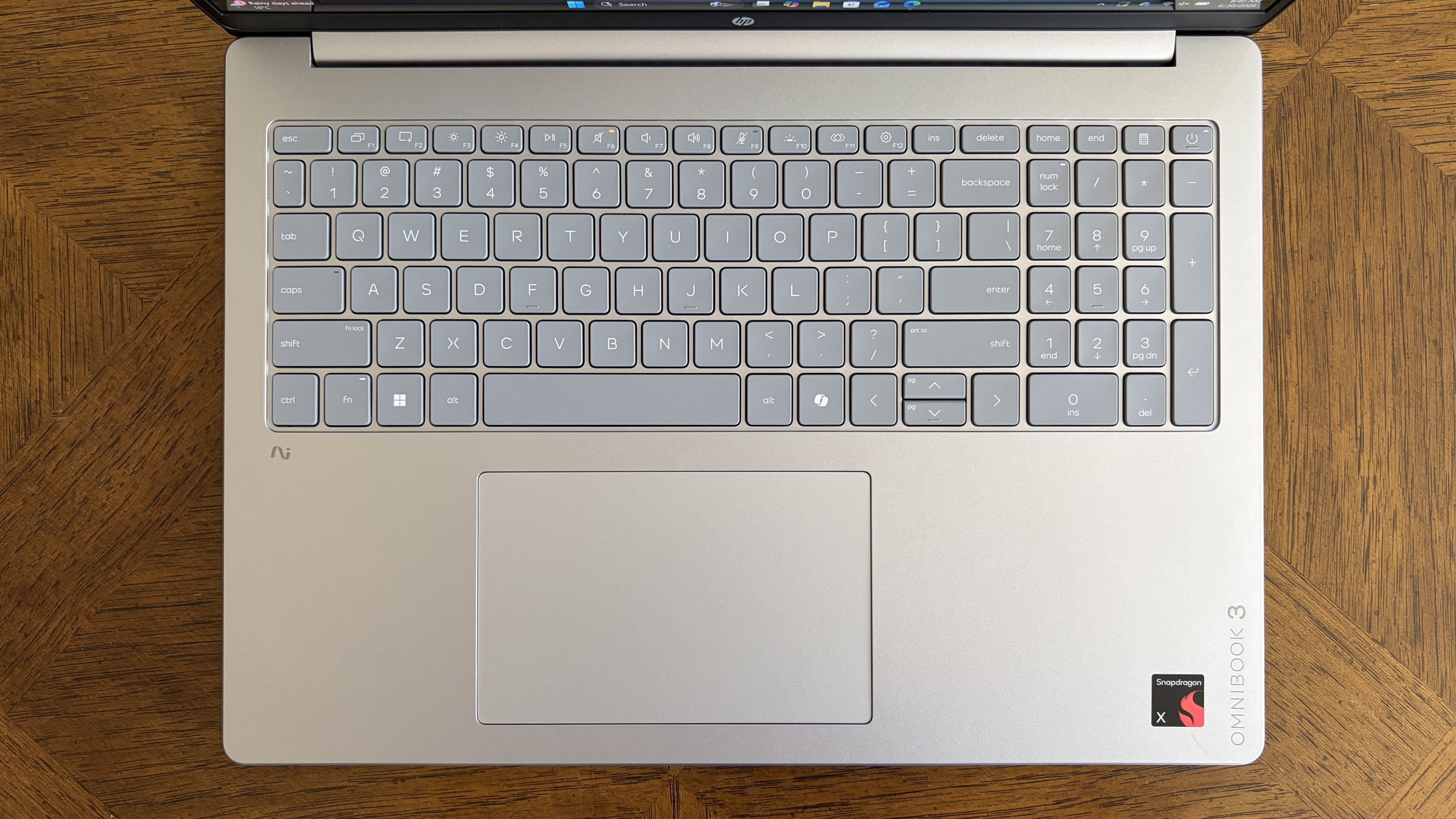 Top view of a silver laptop keyboard with a compact gray key layout and a touchpad, resting on a wooden tabletop. The laptop features &ldquo;OMNIBOOK 3&rdquo; and &ldquo;Snapdragon&rdquo; logos.