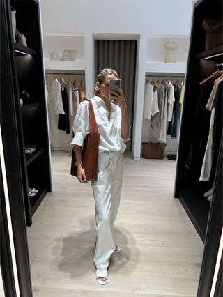 Eliza Huber in the dressing room at Banana Republic wearing a white poplin shirt and white baggy jeans with white flat slides and a brown raffia oversize bag.