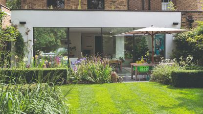 View across the lawn of a shaded garden to the back of the house with the bi-fold doors open. 
