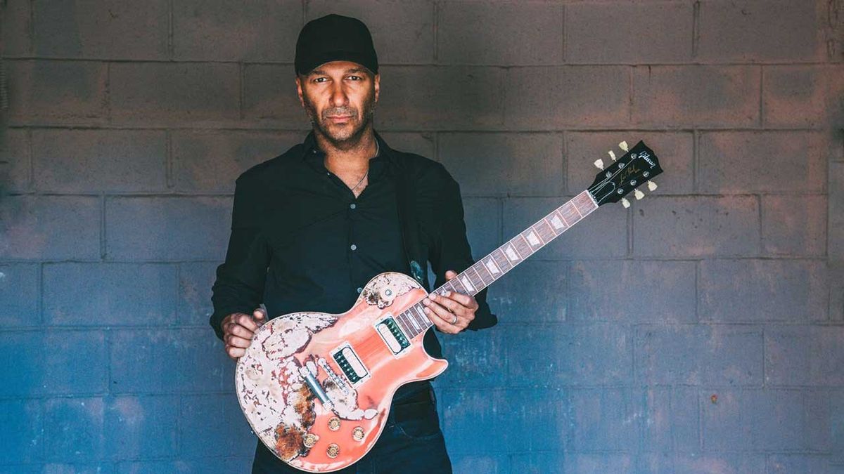 Tom Morello standing against a brick wall holding a guitar