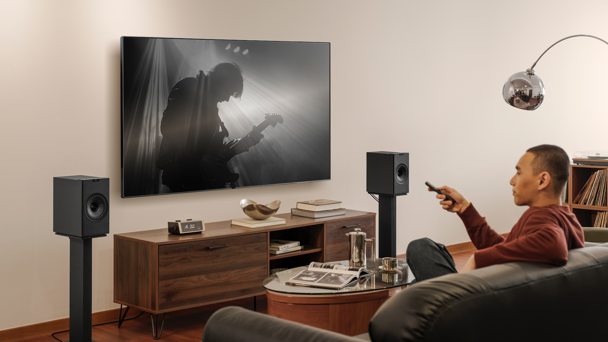 KEF Coda W speakers on stands either side of a TV. A man is watching a music concert on the TV and pointing the remote at it