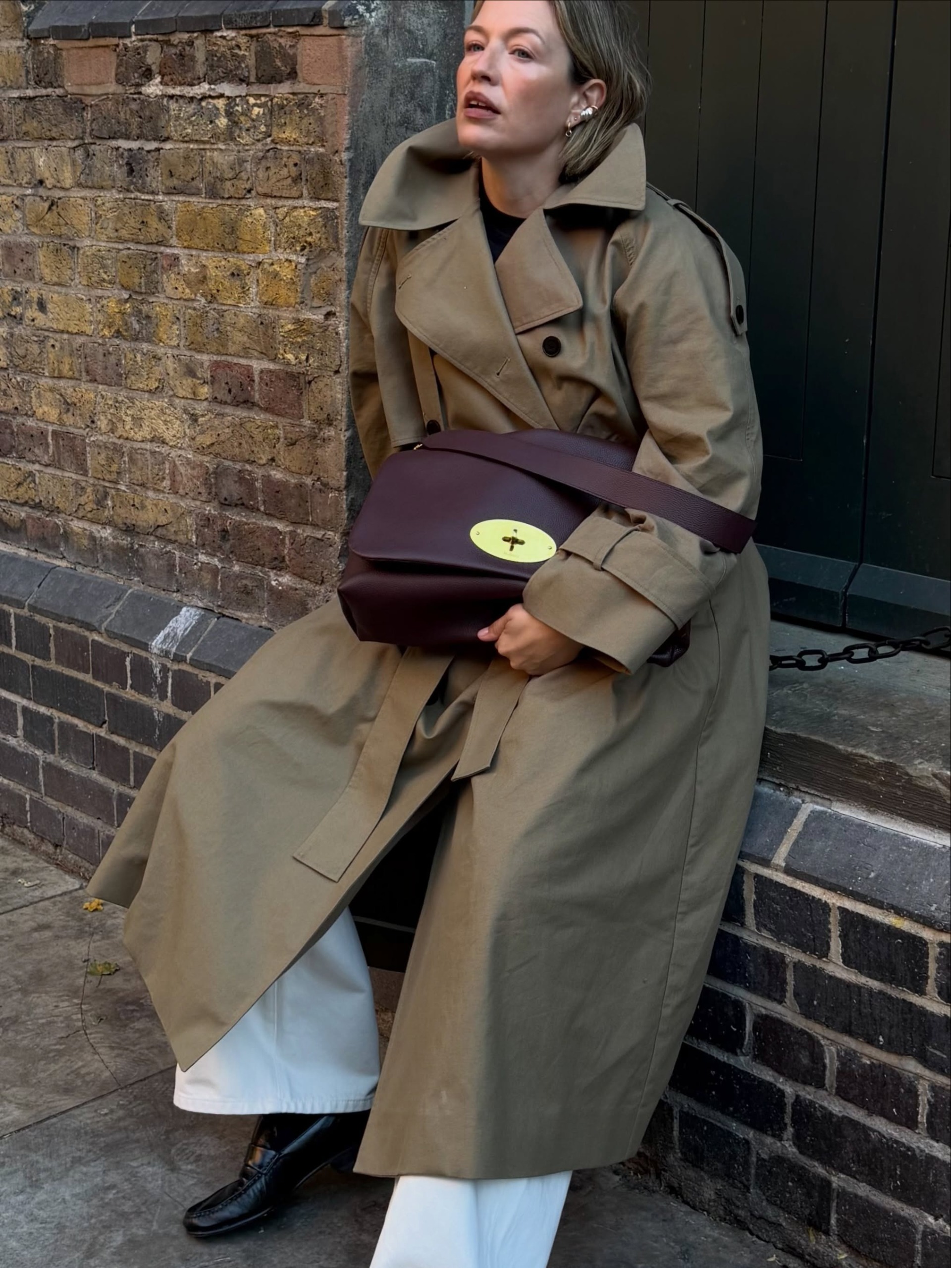 Influencer @alexisforeman poses for a photo wearing a burgundy bag with a trench coat, white jeans and black loafers.