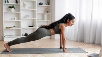 woman wearing khaki crop top and leggings with long hair performing a high plank sideways to the camera and looking at a laptop to the right of her towards us 