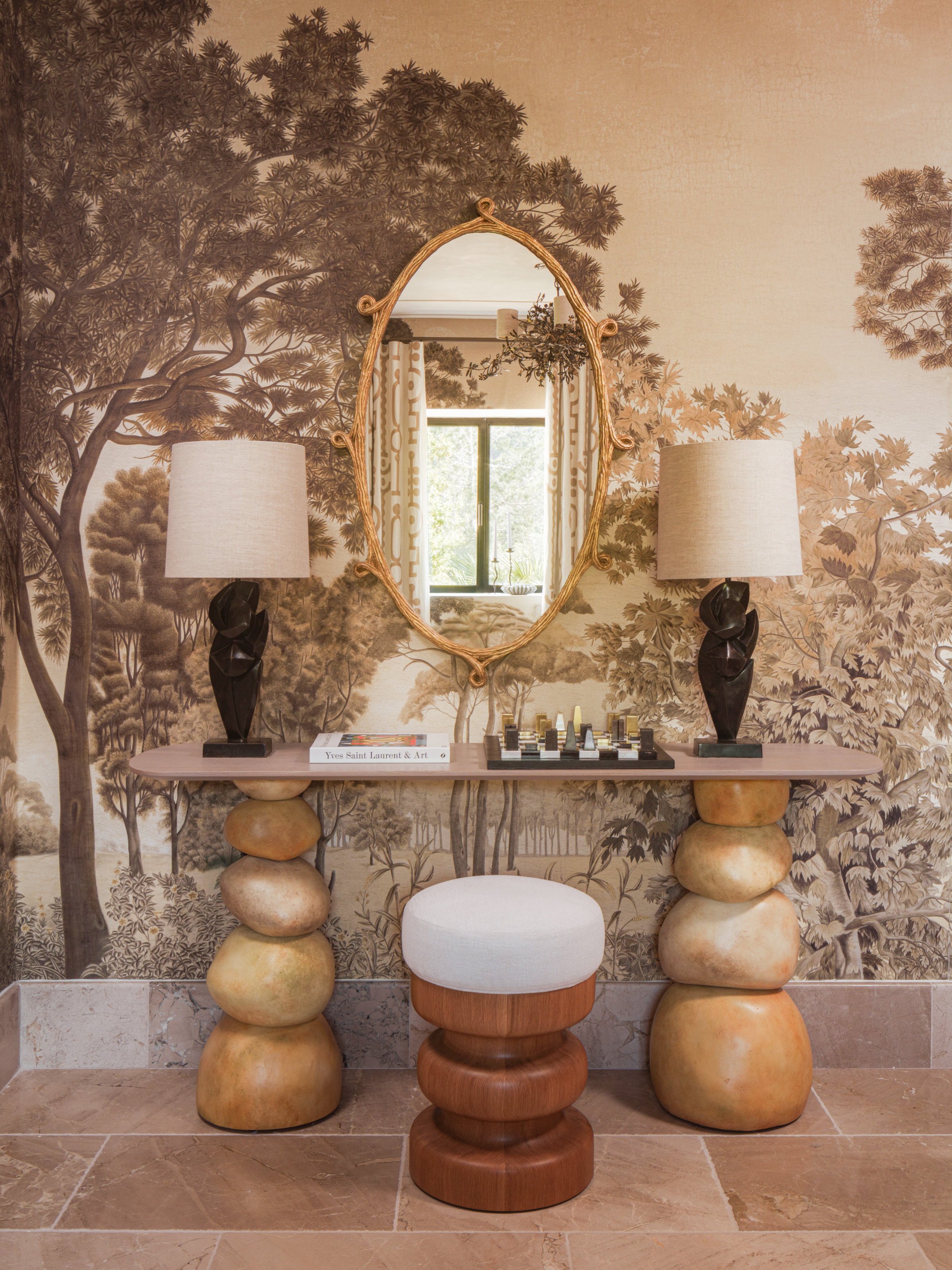 Guest room vanity area with botanical wall mural, sculptural stacked console table, wood and white stool, dark wood lamps framing cane loop framed oval mirror