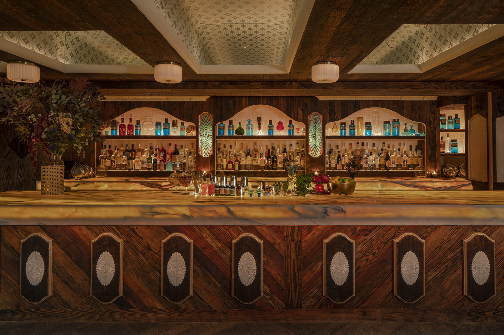A Gatsbyan cocktail bar features marine green details that stand out against sleek woodwork, glass lighting accents, and the colorful selection of liqueurs stored behind the counter.
