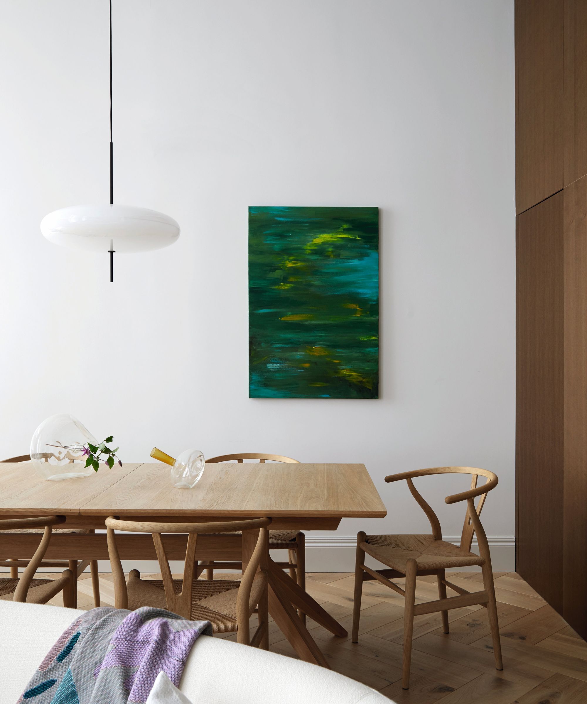 A serene, modern dining space designed by Sandra Flashman, featuring a light wood table, wishbone-style chairs, an abstract green painting on a white wall, and a unique pendant light