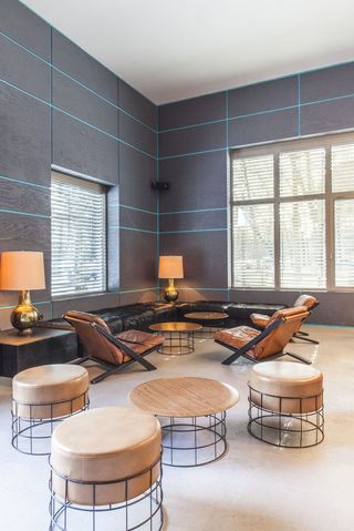 A witting area with round chairs and tables, leather chairs, black coffee and side tables and black leather couches next to a blue striped grey wall.