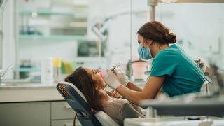 Woman experiencing tongue pain lying down in dentist's chair being treated by dentist with medical face mask on