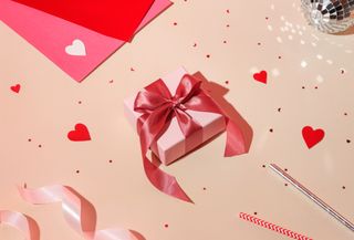 A small, light pink gift box with a pink ribbon, along with red heart confetti and pink paper, on a pastel pink background.