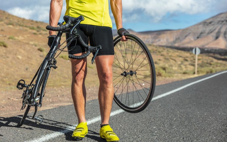In this image of how to fix a puncture is the lower half of a rider holding a front wheel in their right hand and the rest of the bike in their left, while standing on a road with a sandy background. 