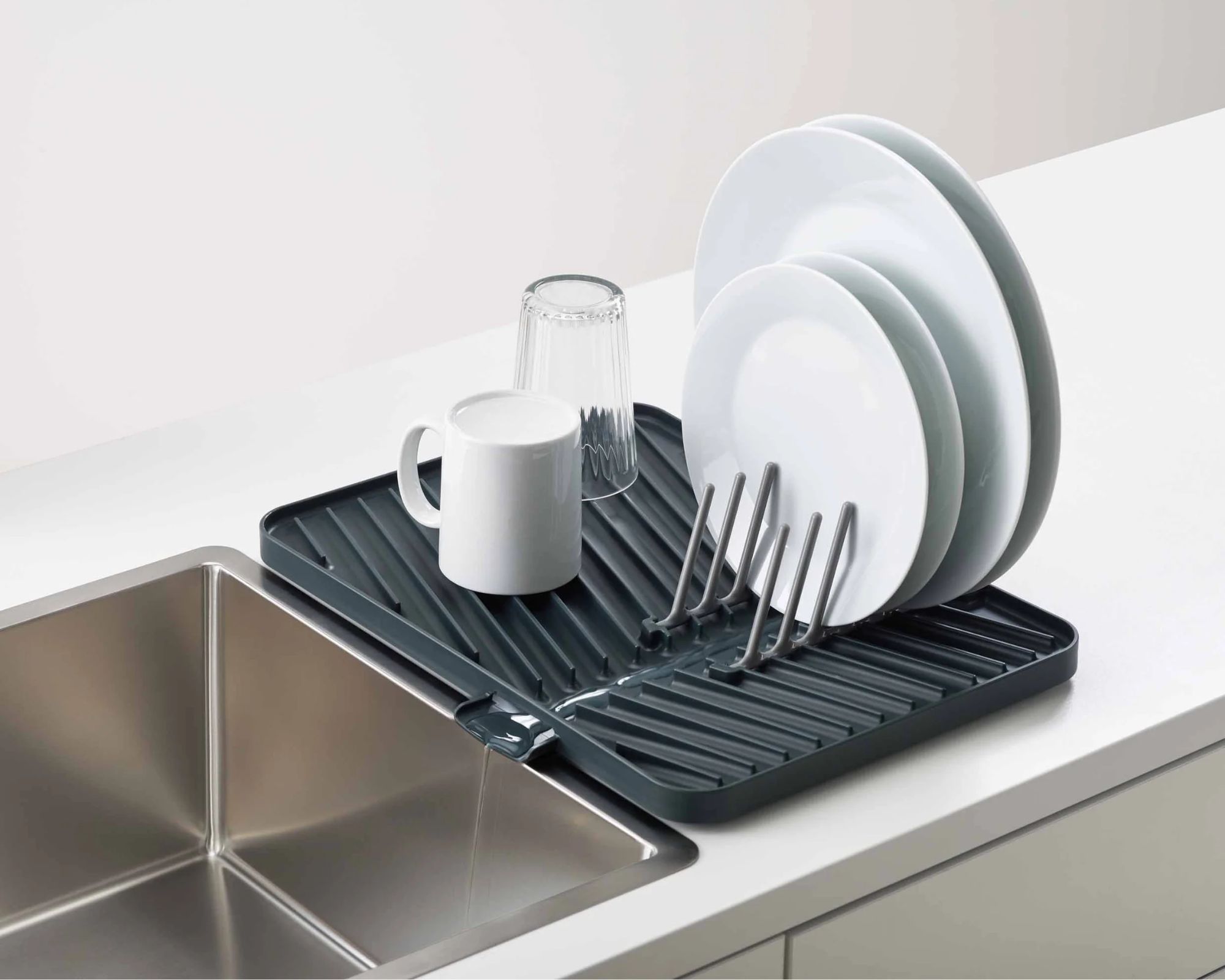 Joseph Joseph flip-up draining board on a white countertop beside a silver stainless steel sink. On the rack are four white plates, a white mug, and a glass.