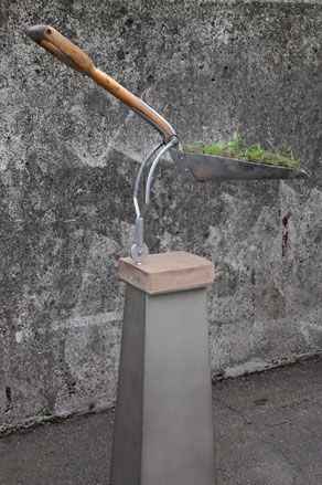 Concrete wall and floor, stone sculpture base with a raised wooden handle spade with a scoop of soil and grass
