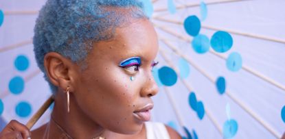 A woman with blue hair and bright makeup underneath an umbrella