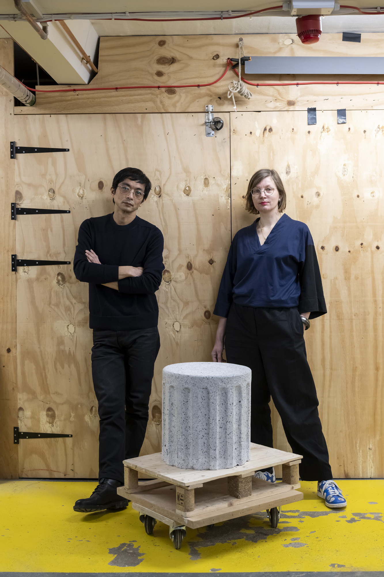 An artist duo dressed in dark workwear tops and trousers and composed of a young woman and a young man with glasses stands against an industrial wooden wall before a stone-like, engraved stool positioned on a wooden trolley.