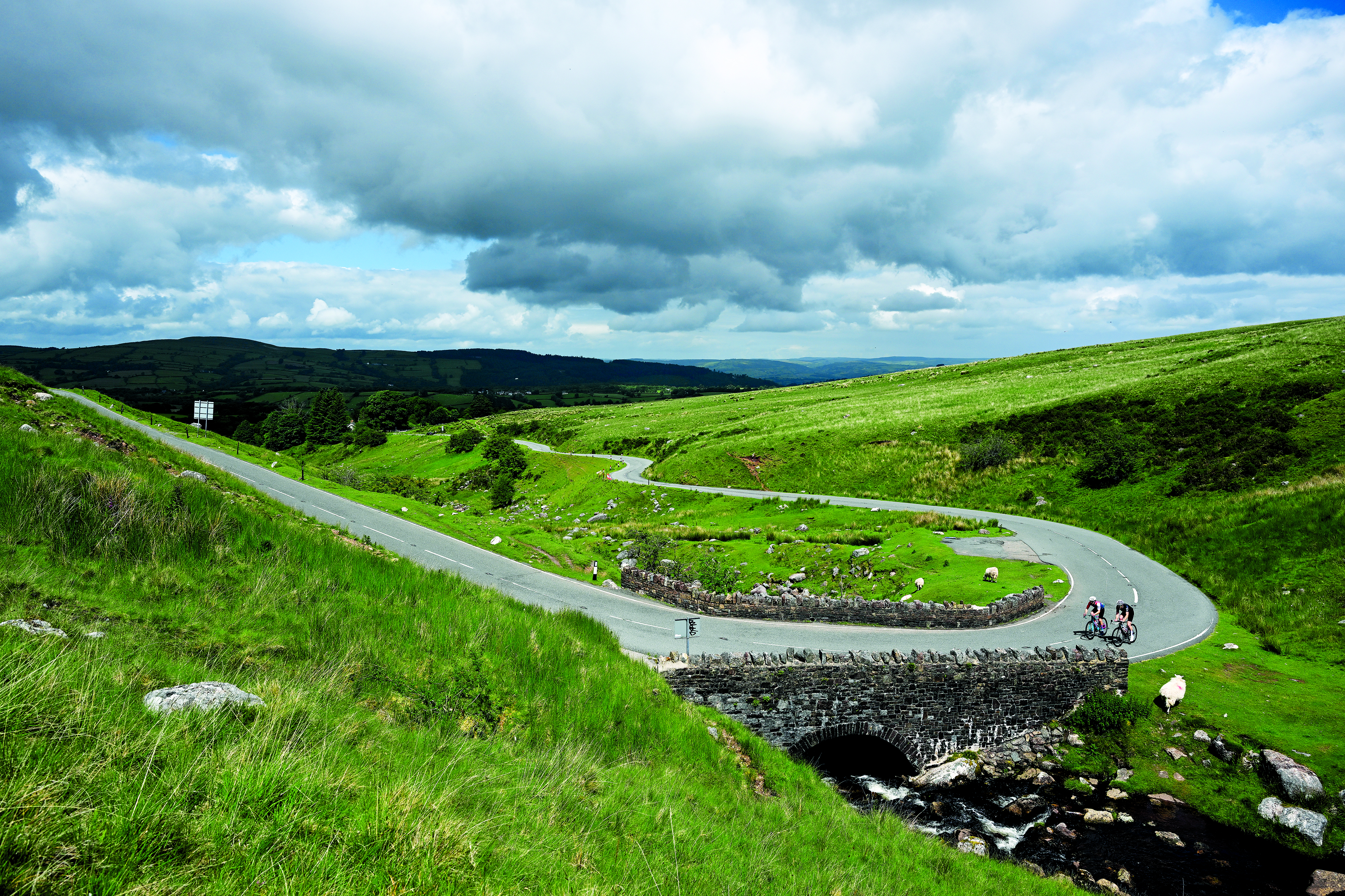 two cyclists ride side by side around a bend surrounded by fields with cloudy skies above them