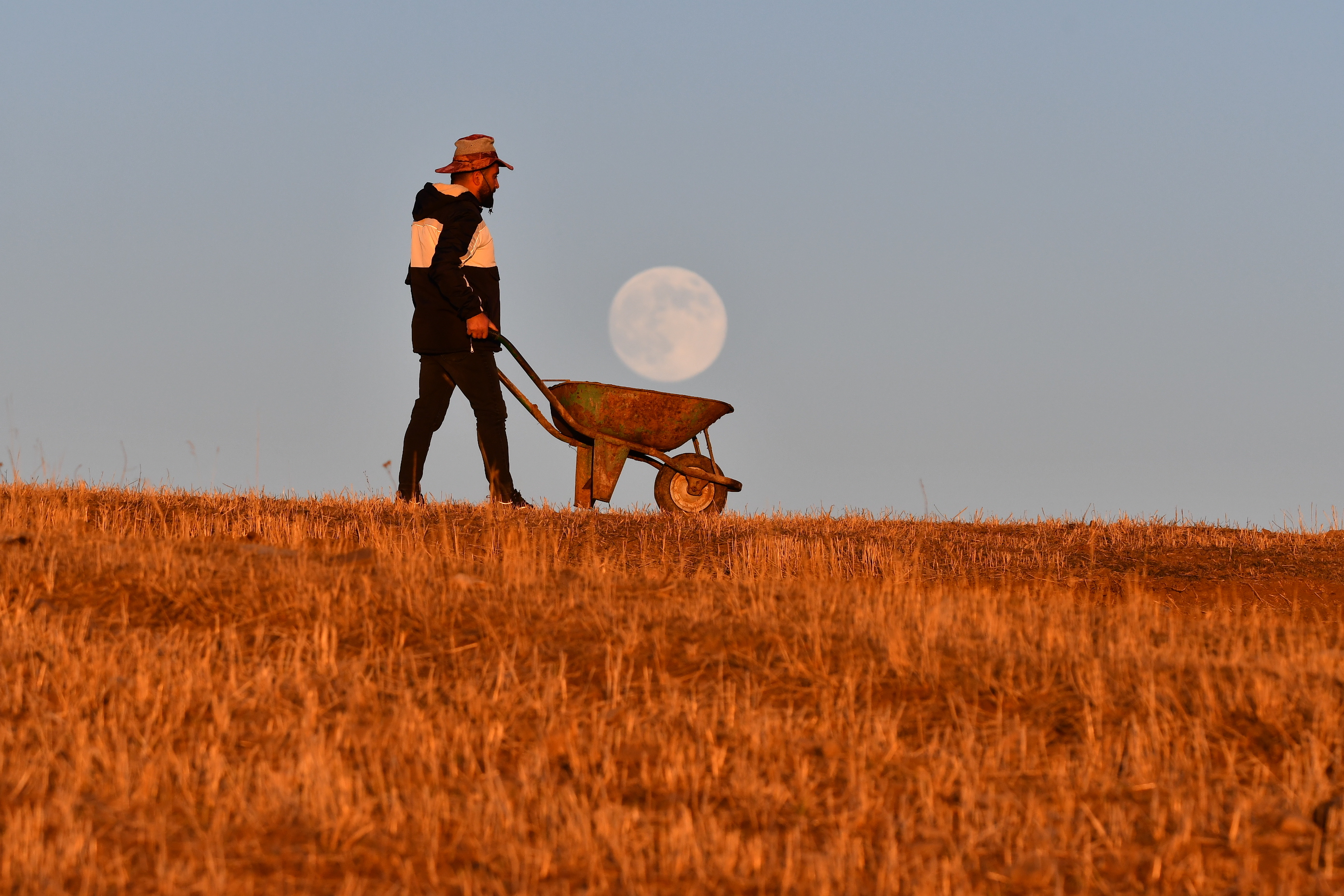 A photo of a man posing with a wheelbarrow as if he&#039;s carrying the moon, which is rising in the background.