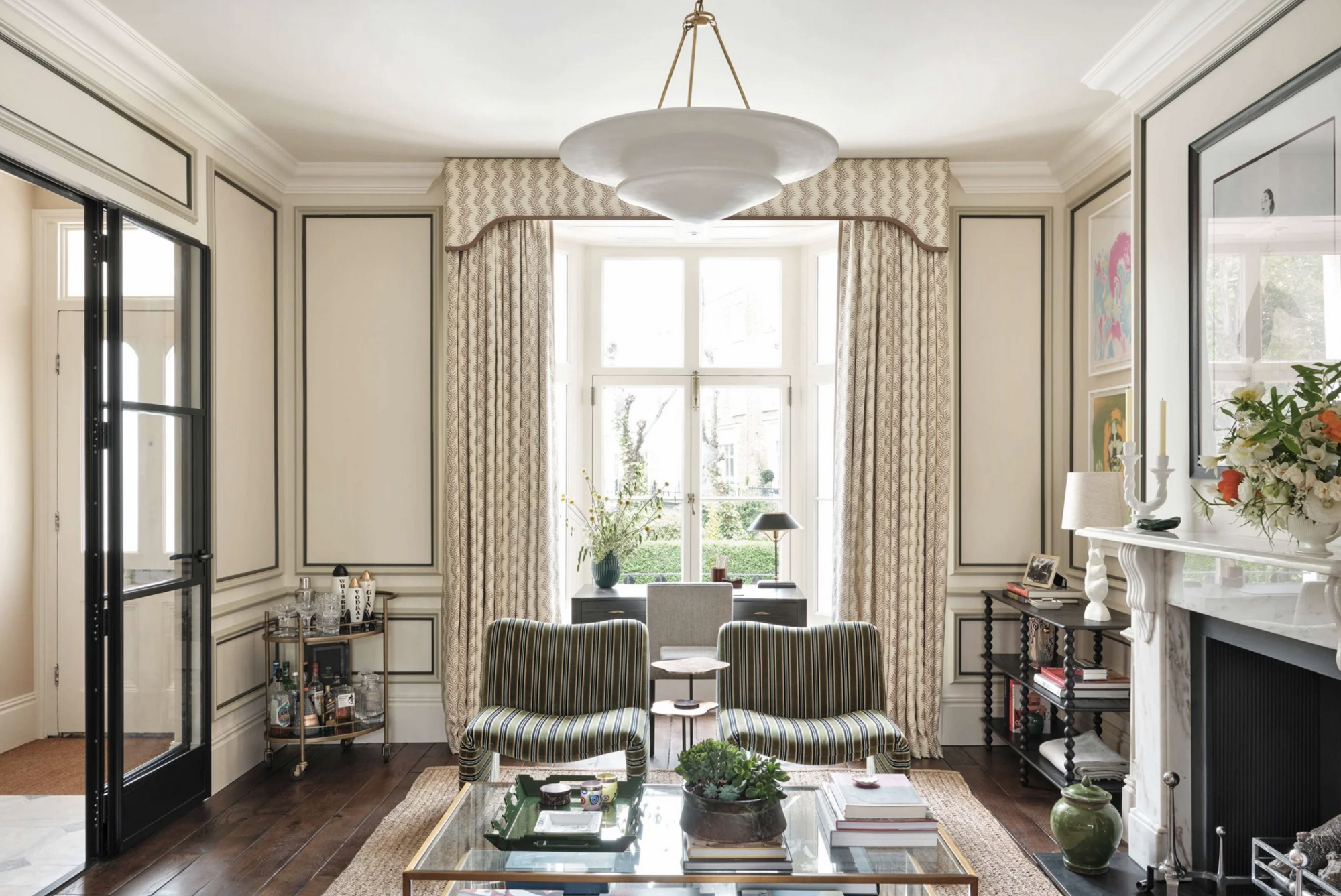 living room with curtain trends with pelmet, moldings, two armchairs, large square glass coffee table styled with coffee table books and vase of flowers, with ornate fireplace