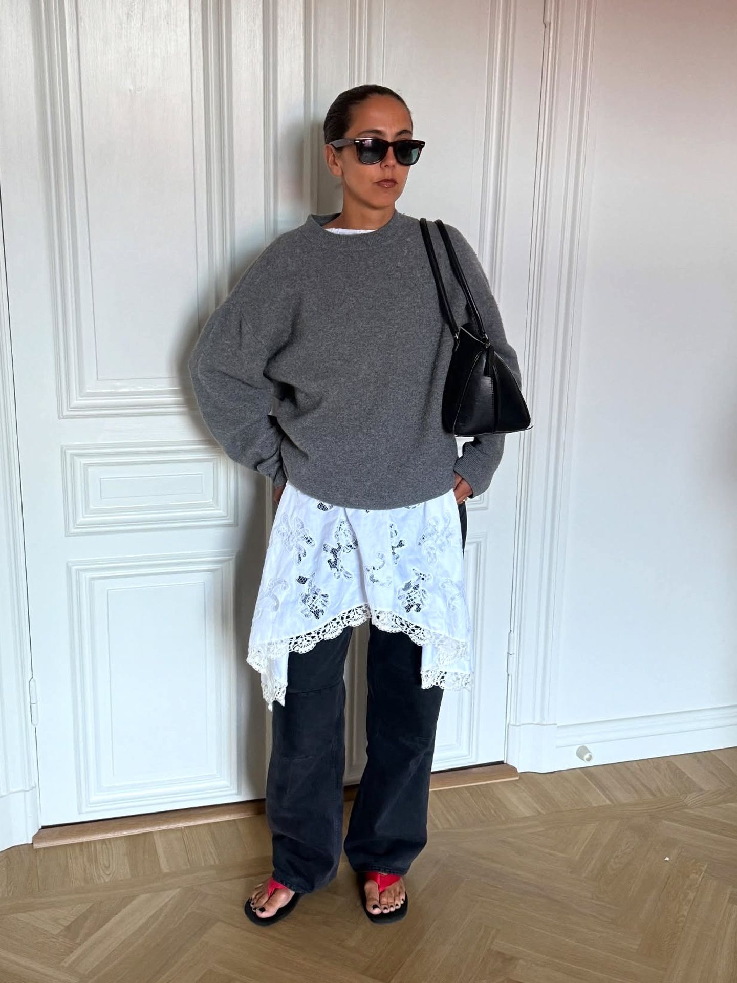 style influencer Hanna MW poses in black sunglasses, an oversize gray sweater layered over a white lace dress, black jeans, and black and red flip-flop sandals