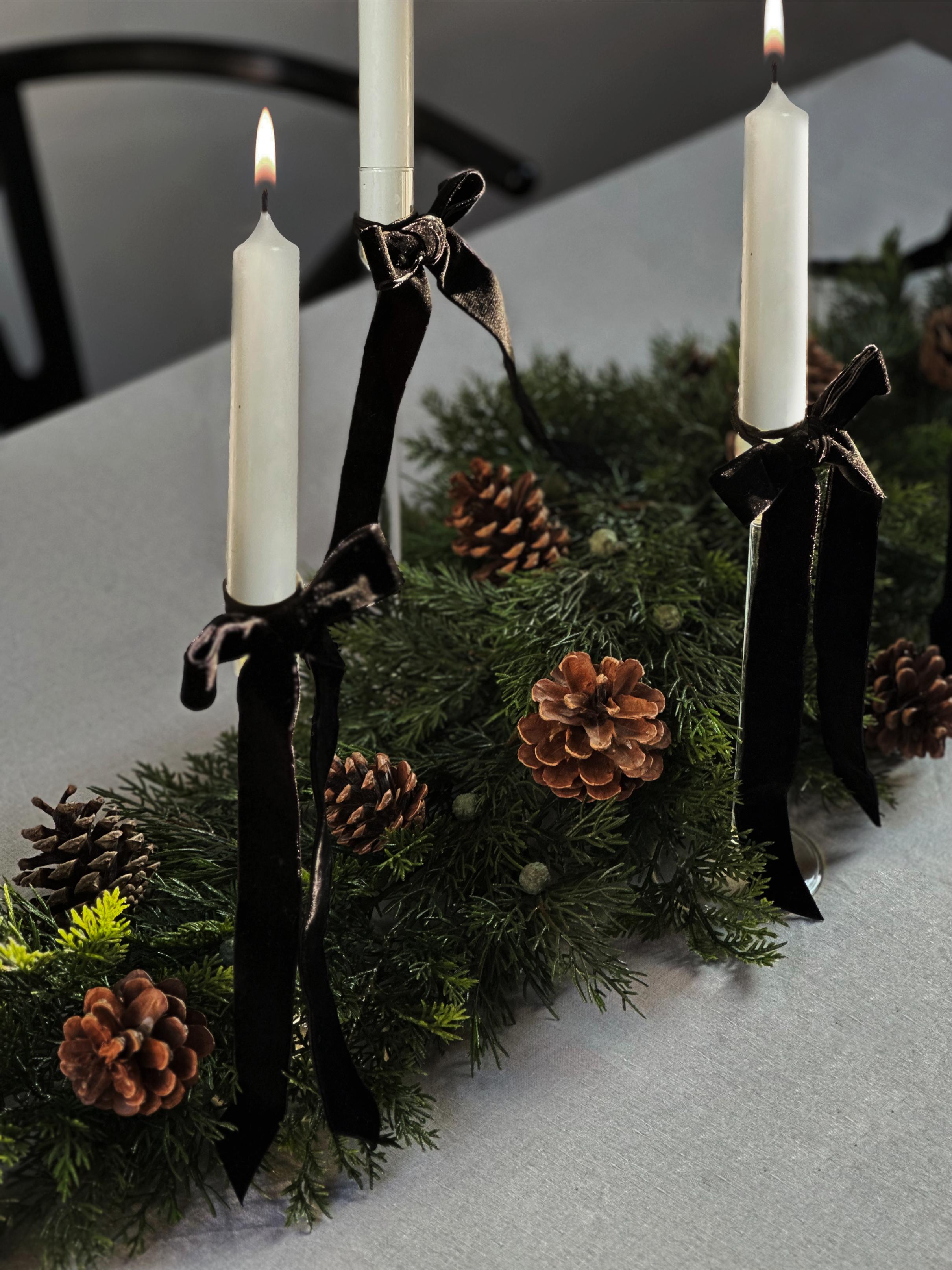 A christmas table decoration on a white tablecloth with evergreen foliage in the center, pinecones, and three candlesticks with white candles wrapped with dark gray bows