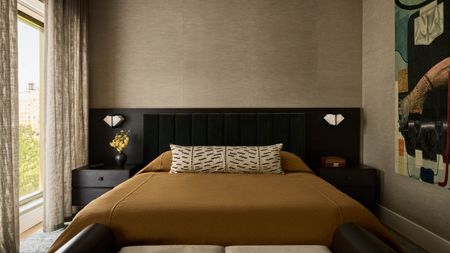 Image of a beige bedroom with a warm color scheme. There is a bed with an extended black headboard and burnt orange bedding. There is a window on the left wall that has semi-sheer, beige, linen curtains.