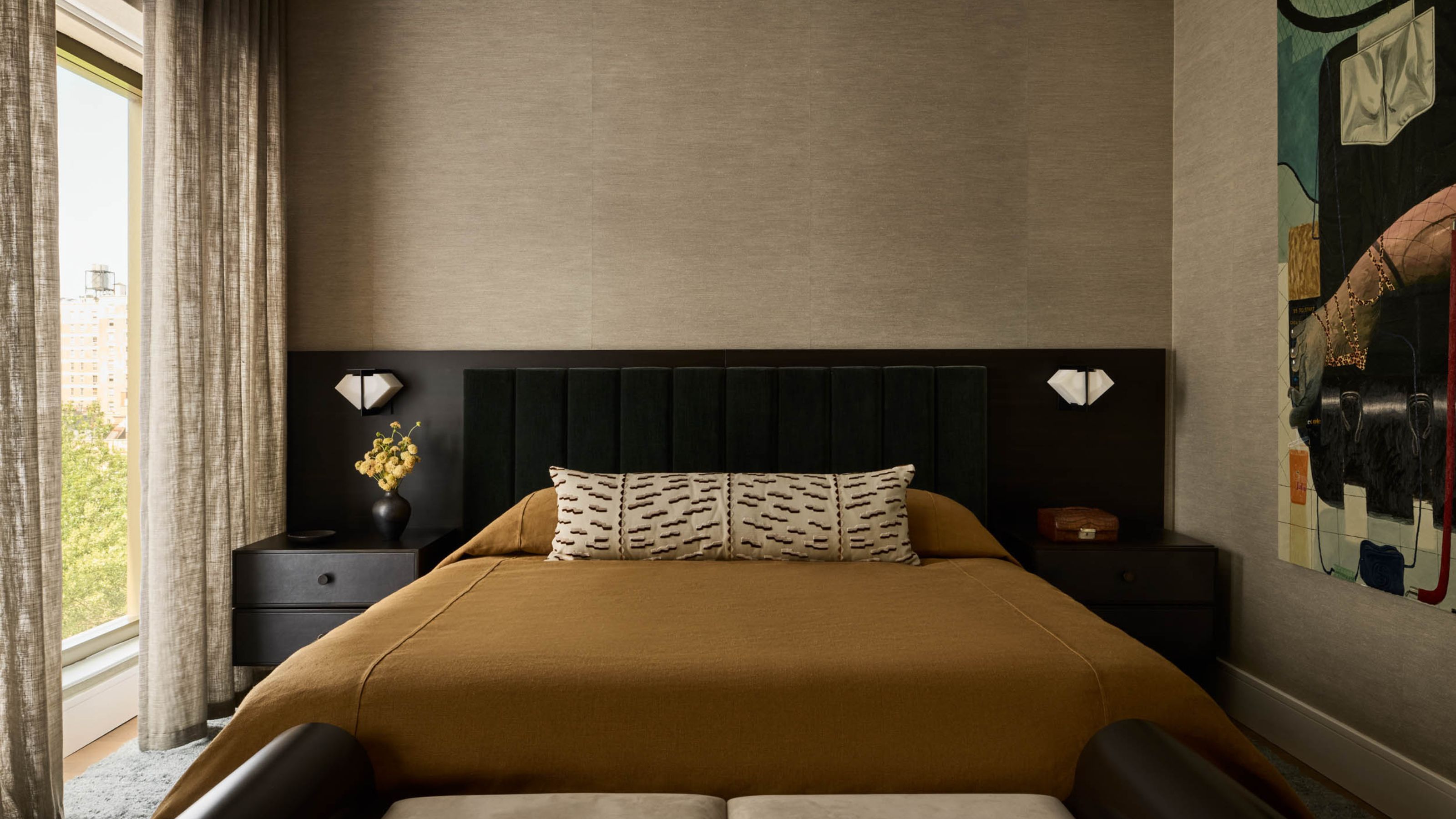 Image of a beige bedroom with a warm color scheme. There is a bed with an extended black headboard and burnt orange bedding. There is a window on the left wall that has semi-sheer, beige, linen curtains.