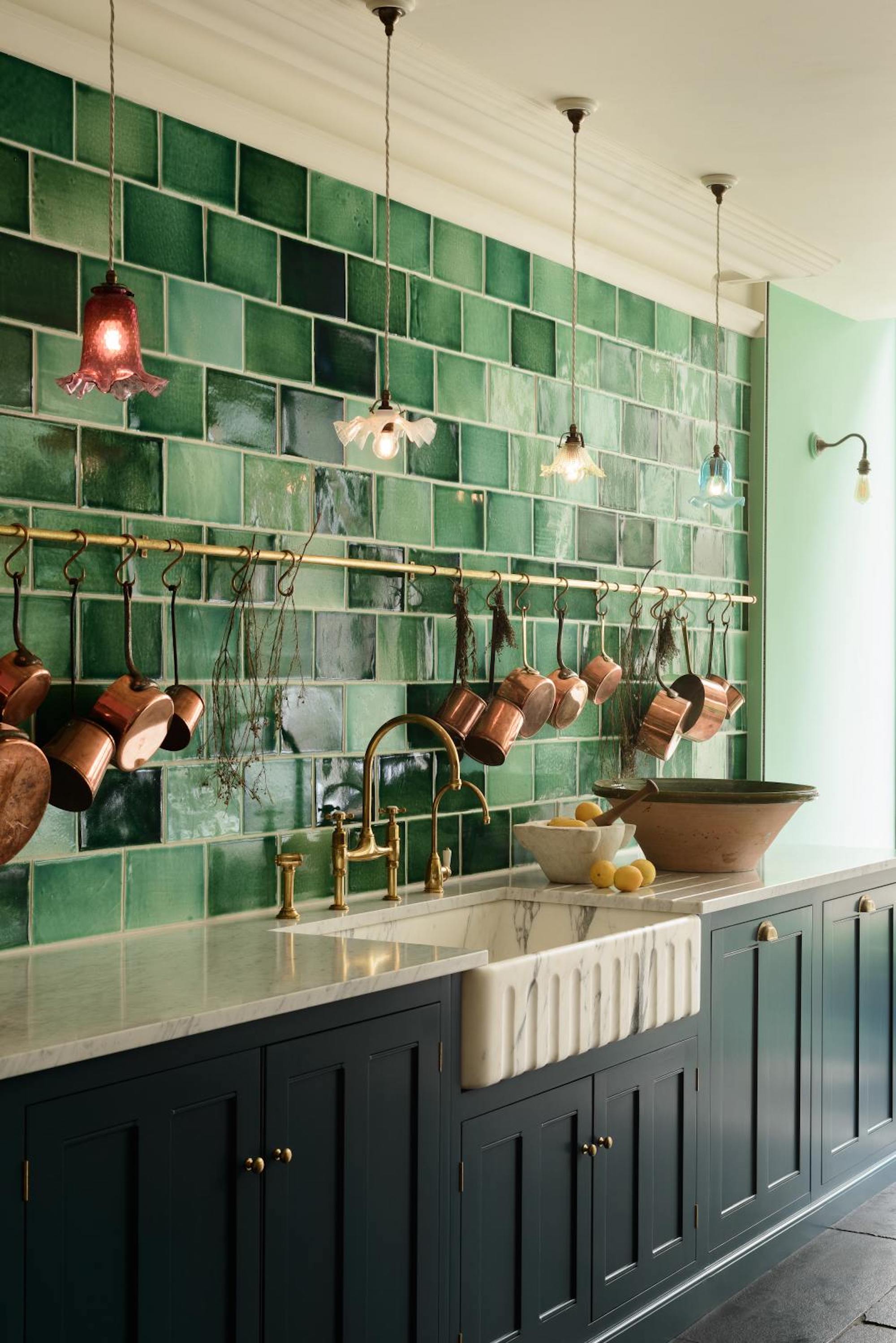 View of the marble kitchen sink, emerald and sage green tiled wall and four pendant lights in front, dark teal cupboards and marble work tops.