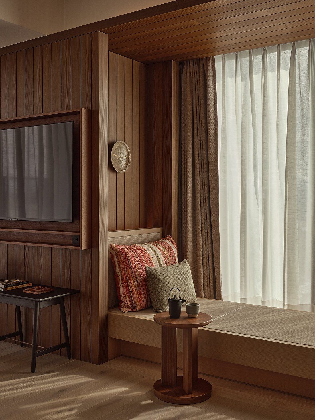 A soothingly decorated hotel suite features wood-paneled walls, earthy furnishings, crafty mugs, coffee tables, and accessories, and natural light filtering through crisp white curtains.
