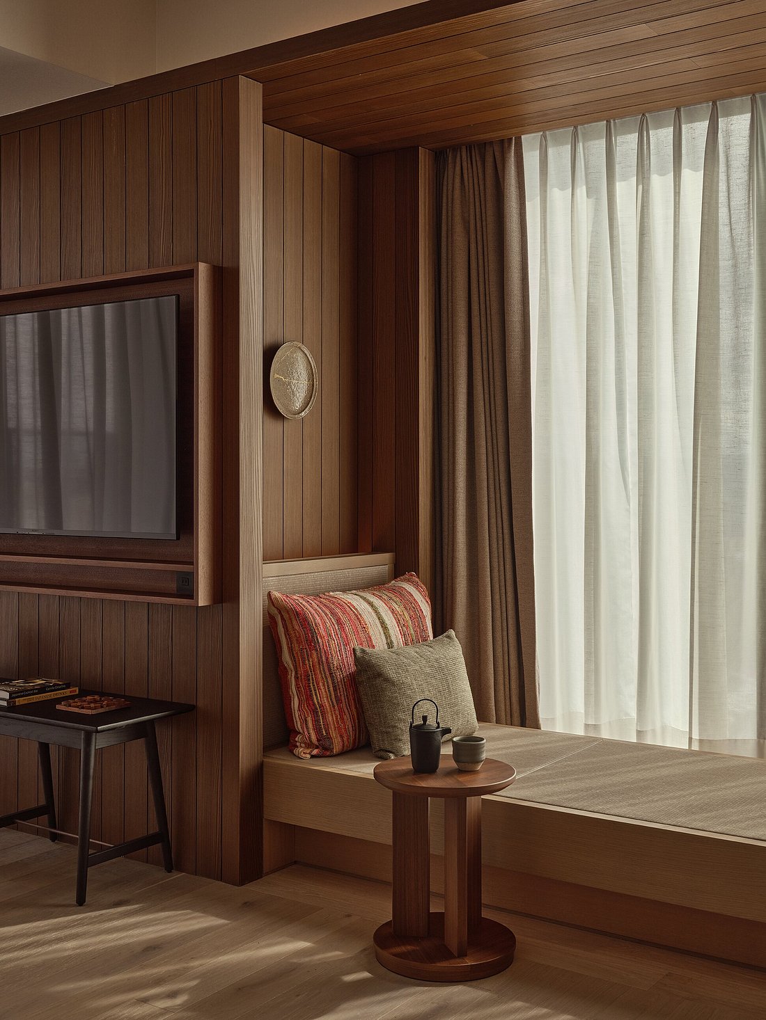 A soothingly decorated hotel suite features wood-paneled walls, earthy furnishings, crafty mugs, coffee tables, and accessories, and natural light filtering through crisp white curtains.