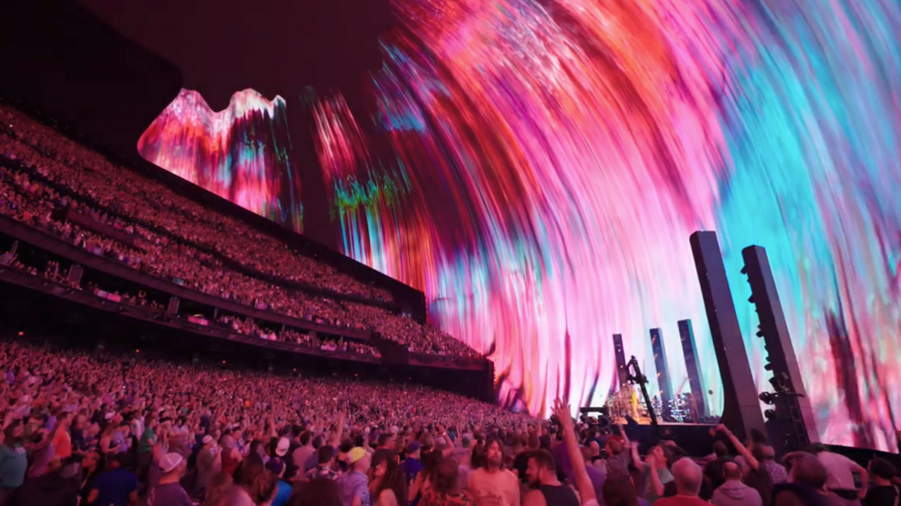 Phish playing on stage in front of a big crowd with a rainbow of colors behind them on the screen at Sphere