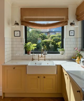 A kitchen with buttery yellow cabinets, white wall tiles, and natural hessian-toned window treatments