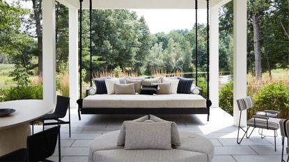 outdoor living room with garden swing chair, dining table, round ottaman-style seat, and armchairs, with views of verdant gardens wrapping the area