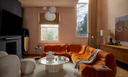 Living room with plaster pink walls, black plaster finish fireplace wall, orange corner sofa, white stone coffee table, beige armchair and white ceiling lights