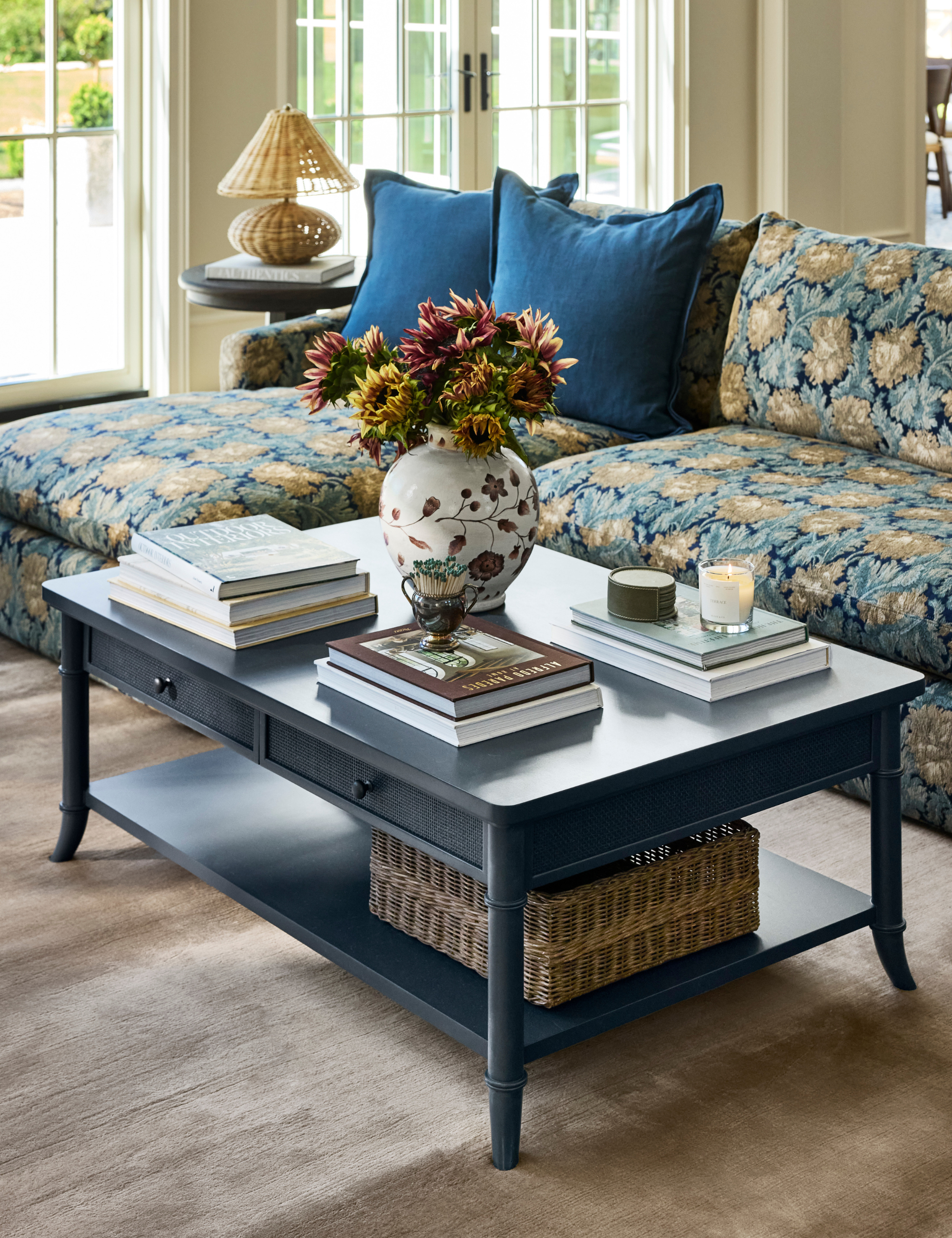 Living room featuring a floral jacquard sectional. two-tier navy storage coffee table, taupe rug, woven table lamp, and collected objects
