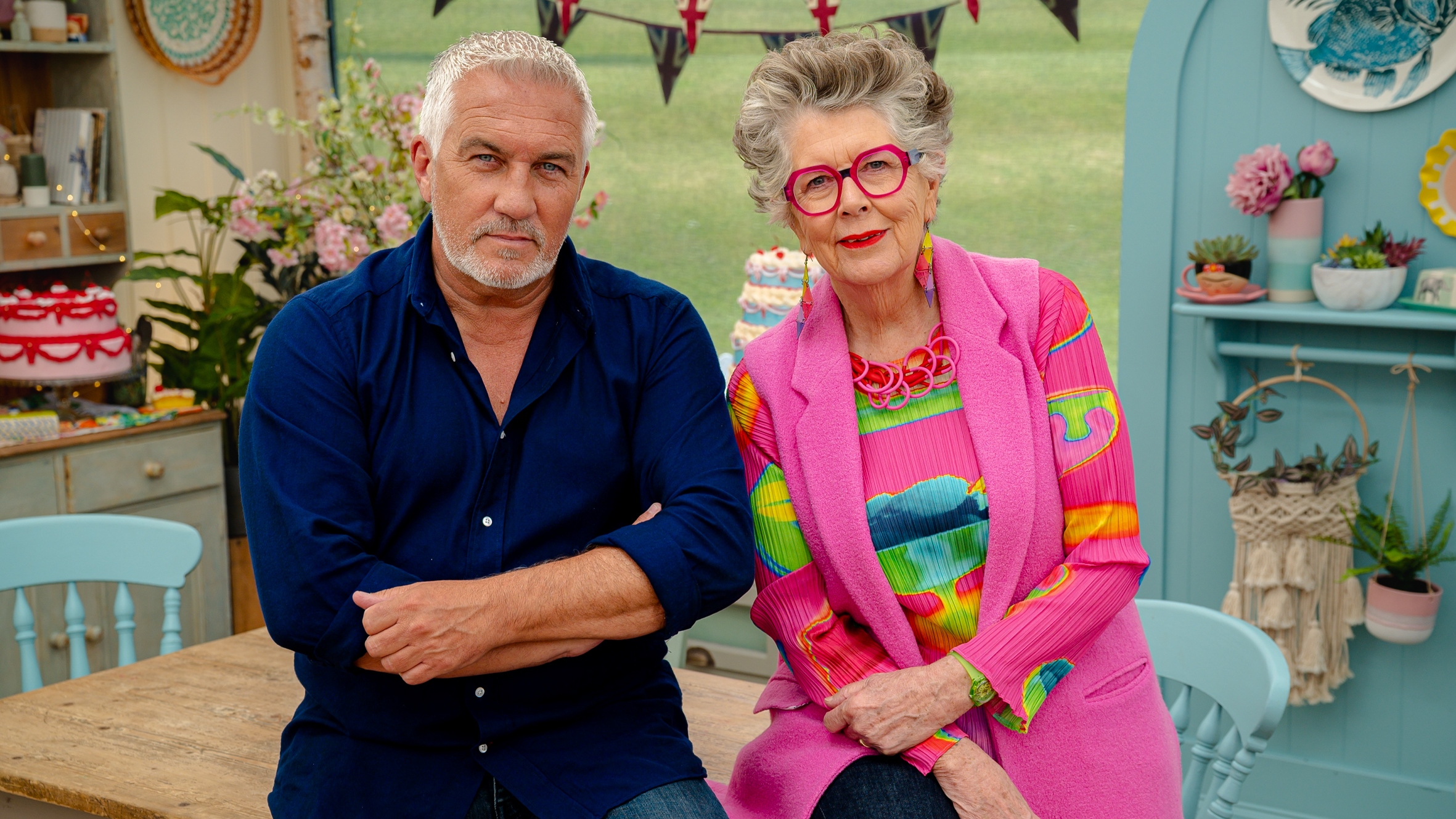 Paul Hollywood in a dark shirt and Prue Leith in a pink tunic and patterned top in the tent in The Great British Bake Off 2025