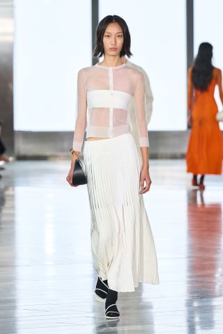 A photo from the Jonathan Simkhai S/S 25 runway show of a model wearing a sheer white cardigan and plissé skirt with black tights and strappy sandals.