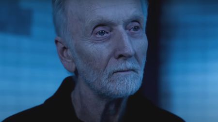 Tobin Bell smiles slightly in a blue lit torture room in Saw X.