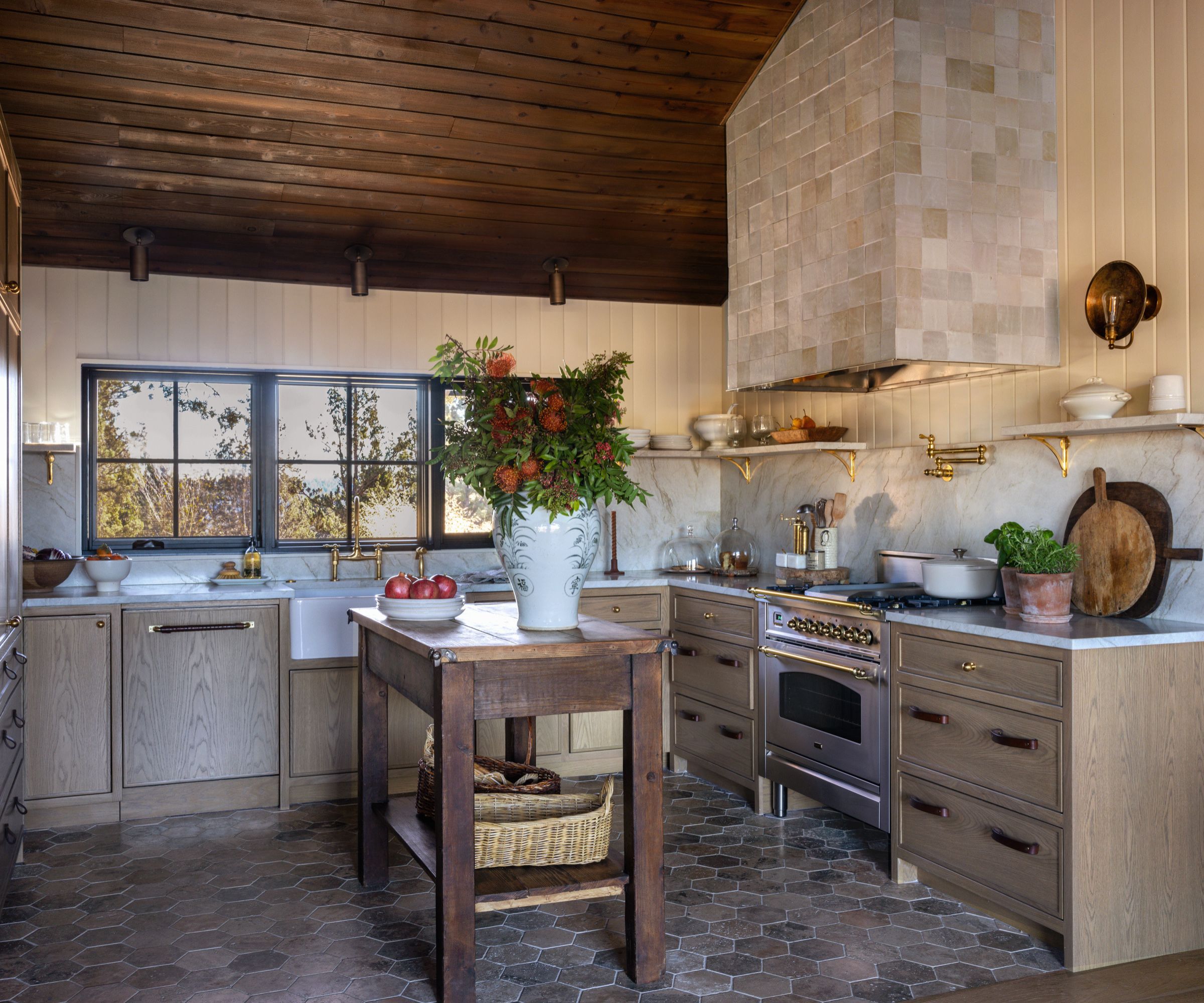 A kitchen with wood cabinetry, open shelves, and brass fixtures. The backsplash is marble, and the stove sits under a zellige tiled range hood. A small wooden island with a floral arrangement is in the middle of the space