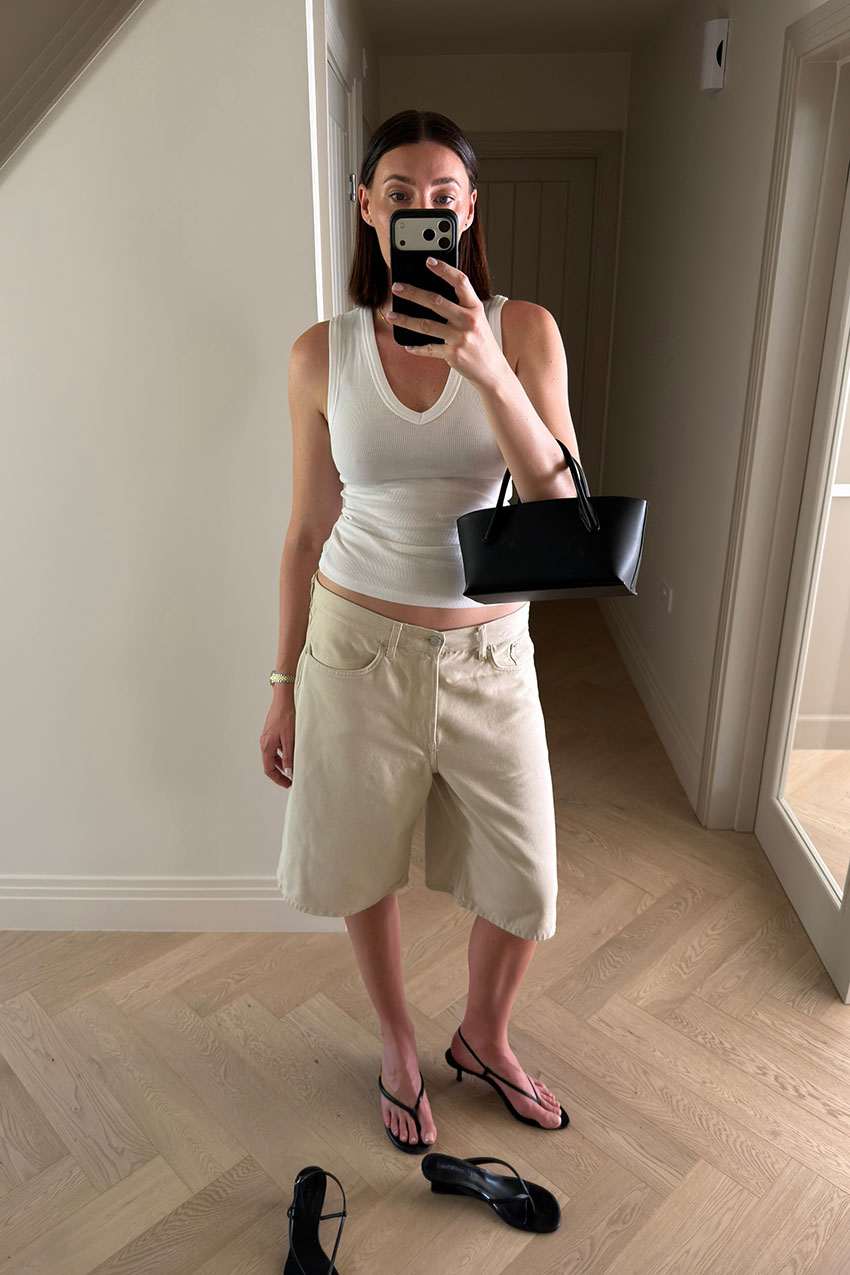 a woman wearing a white V-neck tank top, cream long shorts, black slingback thong sandals and a black east west bag