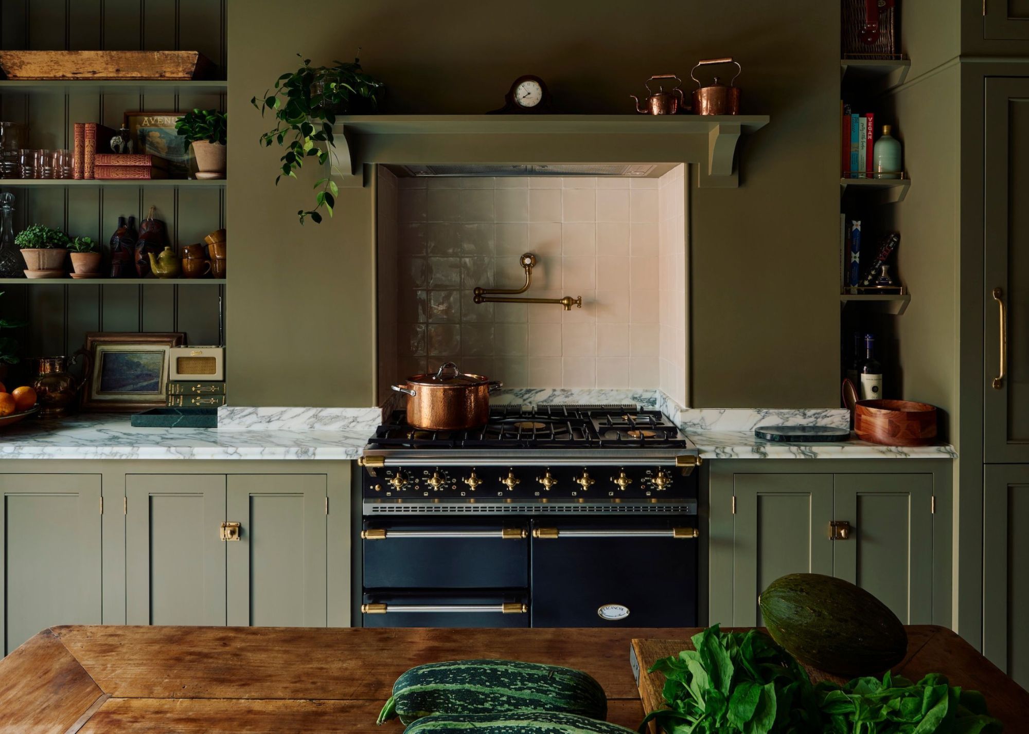 A taditional green kitchen with marble counter, open shelving, aga stove, timber island bench with vegetables on it