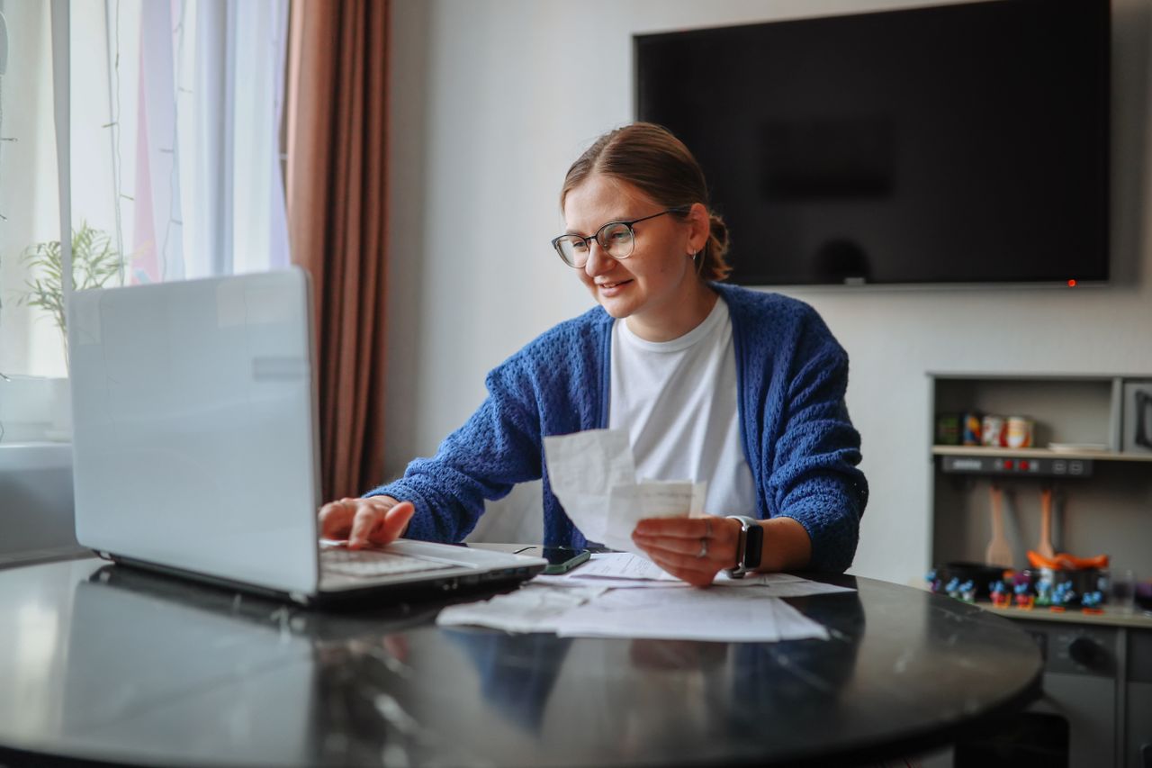 Woman holds receipts as she looks at laptop at table.