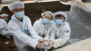 Archaeologist Li Nan (center) with two members of Peking University's archaeological team at the Sanxingdui archaeological site in China's Sichuan province. (plus one more person working in the back). Everyone is wearing hair covers, face masks, and white protective suits.