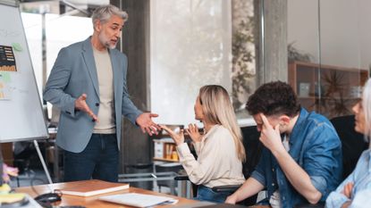 Male and female business executives engaged in a heated argument during a boardroom meeting, with colleagues sitting uncomfortably with heads in hands.