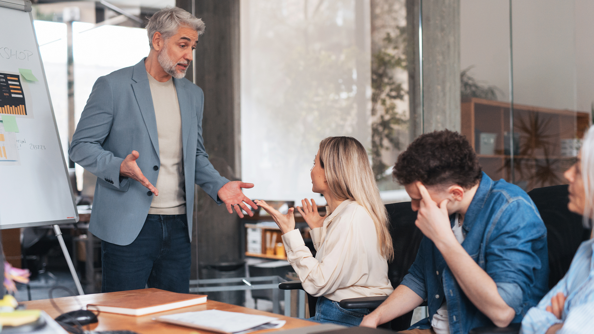Male and female business executives engaged in a heated argument during a boardroom meeting, with colleagues sitting uncomfortably with heads in hands. 