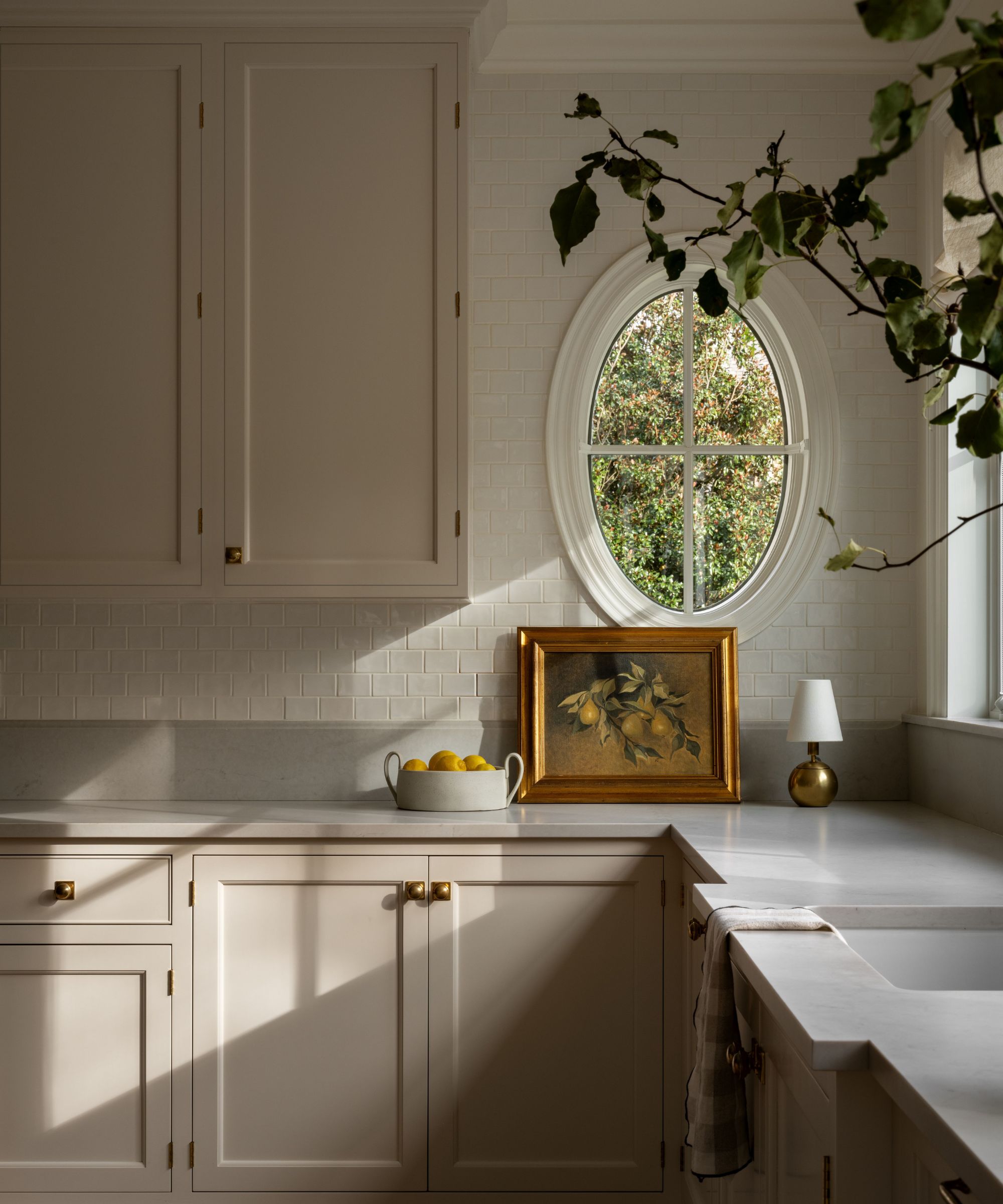 The corner of a warm neutral kitchen with an oval window, a small brass lamp, and a still life lemon painting in a brass frame